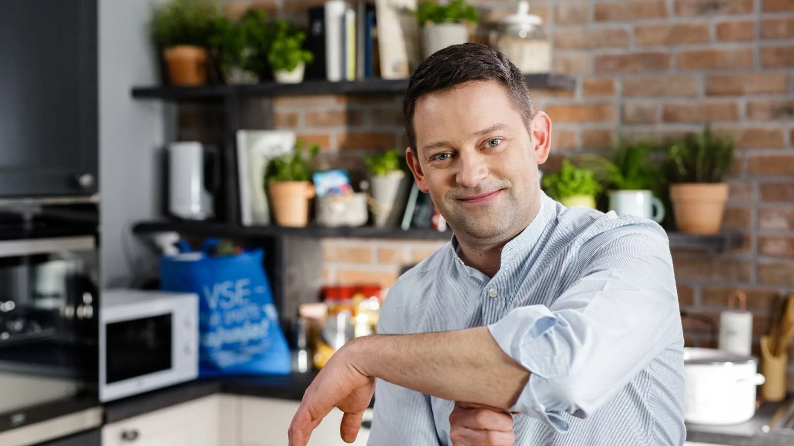 Cooking show host Grega Rozman leans casually on a kitchen counter, smiling at camera, with herb-lined shelves and exposed brick visible behind him